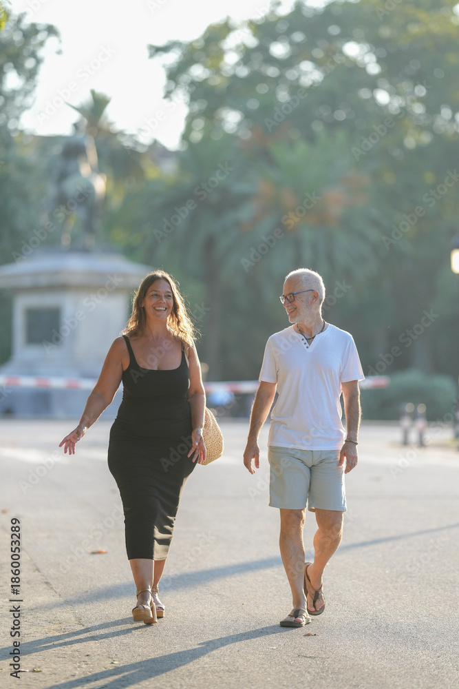 Fototapeta premium A man and a woman are walking down a street. The man is wearing a white shirt and shorts, while the woman is wearing a black dress. They are both smiling and seem to be enjoying their walk