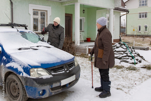 Elderly woman with walking stick talking to an adult man clearing snow from a car. Winter weather and car maintenance concept. Life in country house.