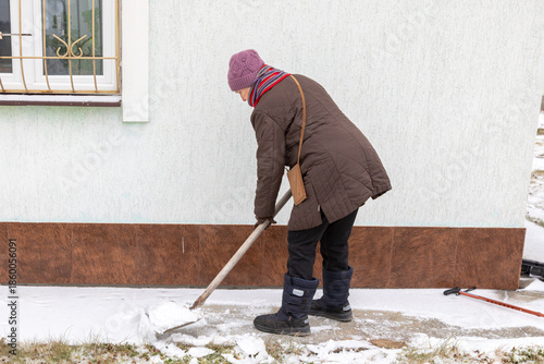 Elderly woman clearing snow with a shovel from the path near her country house wall. Winter household chore concept.