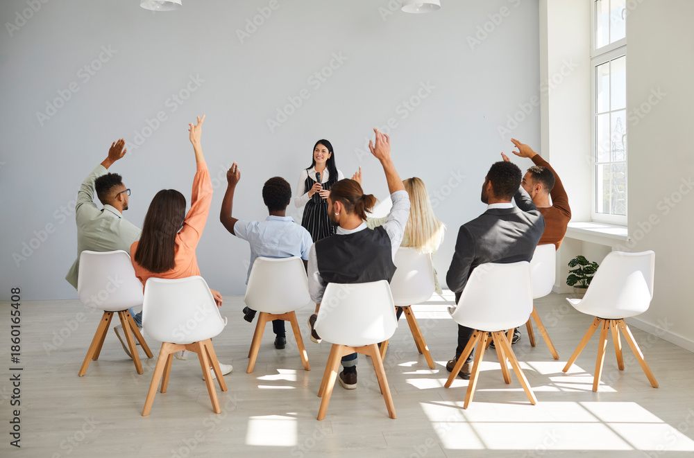 Fototapeta premium Group audience seminar training meeting presenter speaker. Professionals sit in office chairs, raising hands to ask questions during corporate training. Team learning and communication concept.