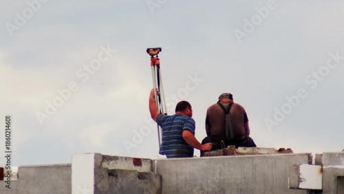 Two construction workers on high concrete wall of building site, one man standing and holding tripod with surveying device, another worker sitting nearby, working on outdoor project
