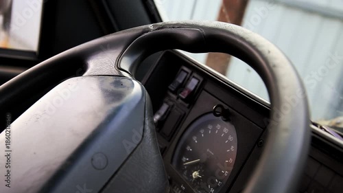 Close up view of steering wheel and control panel from old vehicle. Dashboard features speedometer gauge and various buttons. Worn texture of steering wheel material visible.