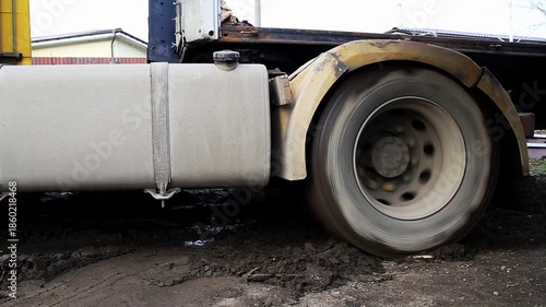 Truck wheel spins rapidly in thick mud. Struggle for traction. Mud churns, splatters. Scene shows vehicle difficulty on unpaved terrain. Outdoors, daytime.