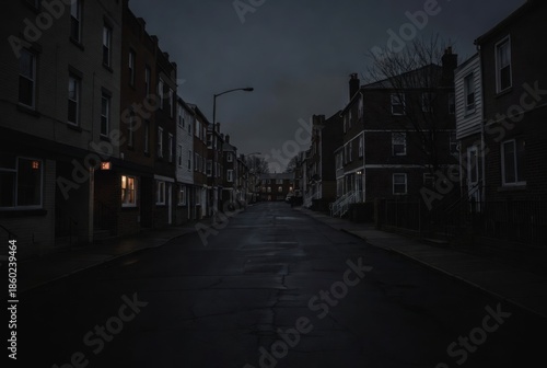 Dark and moody street view of a quiet residential neighborhood at dusk; empty asphalt road lined with traditional brick row houses under a gloomy overcast sky.