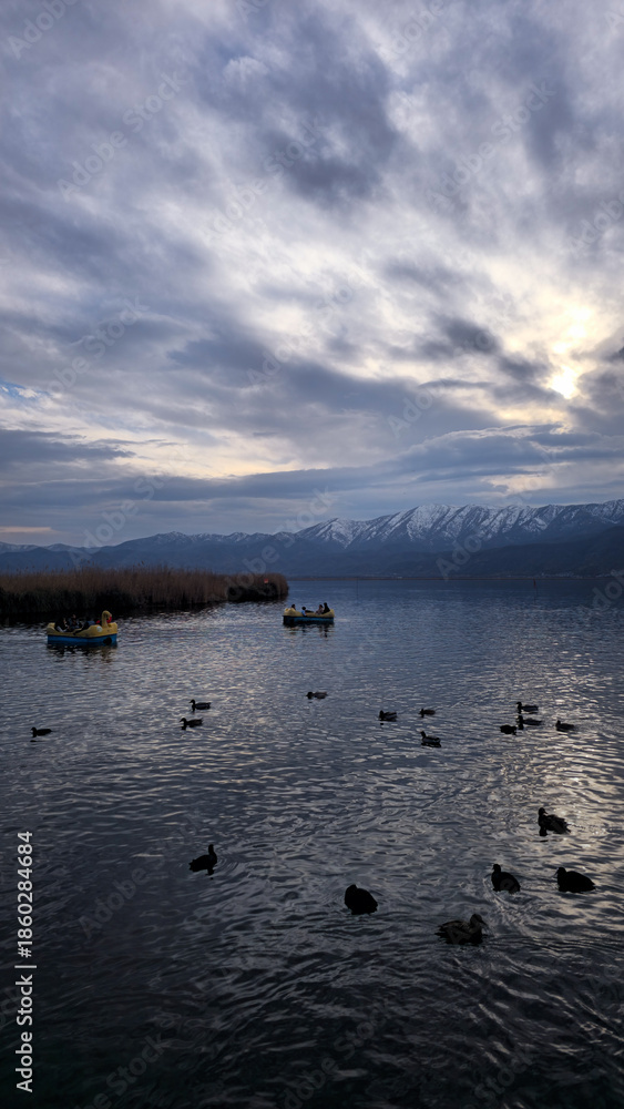 Fototapeta premium Winter scene at Zarivar Lake with ducks on calm water, small boats near reed beds, distant mountains, and soft cloudy light creating a peaceful natural landscape. 📍Marivan, Kurdistan 