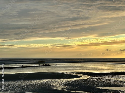 Silhouetted walkers on sandbar at low tide in Authie Bay near Berck, France, golden sunset sky over tidal flats with distant seals resting on sandbanks