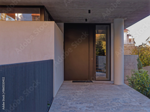 A modern villa entrance with a dark minimalist door and grey-veined stone paving. Brutalist concrete architecture and sleek lines define a high-end, contemporary luxury residence.