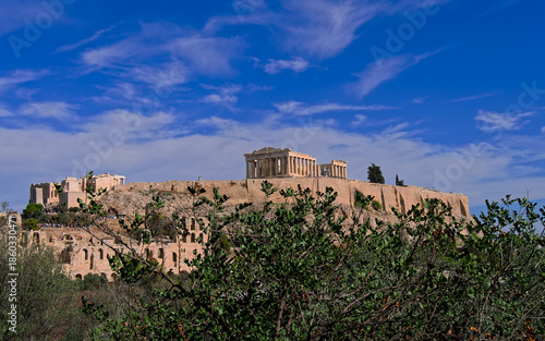 Panoramic view of Ancient Acropolis of Athens and the majestic Parthenon, a Greek cultural landmark, framed by vibrant  foliage in foreground beneath a deep, dynamic blue sky with scattered clouds.