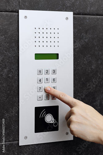 Close-up of a hand pressing a button on a modern intercom keypad mounted on a dark textured wall.