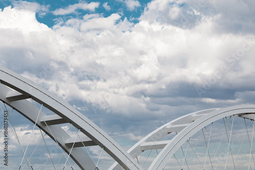 Abstract view of steel bridge arches and suspension cables against a dramatic cloudy blue sky.