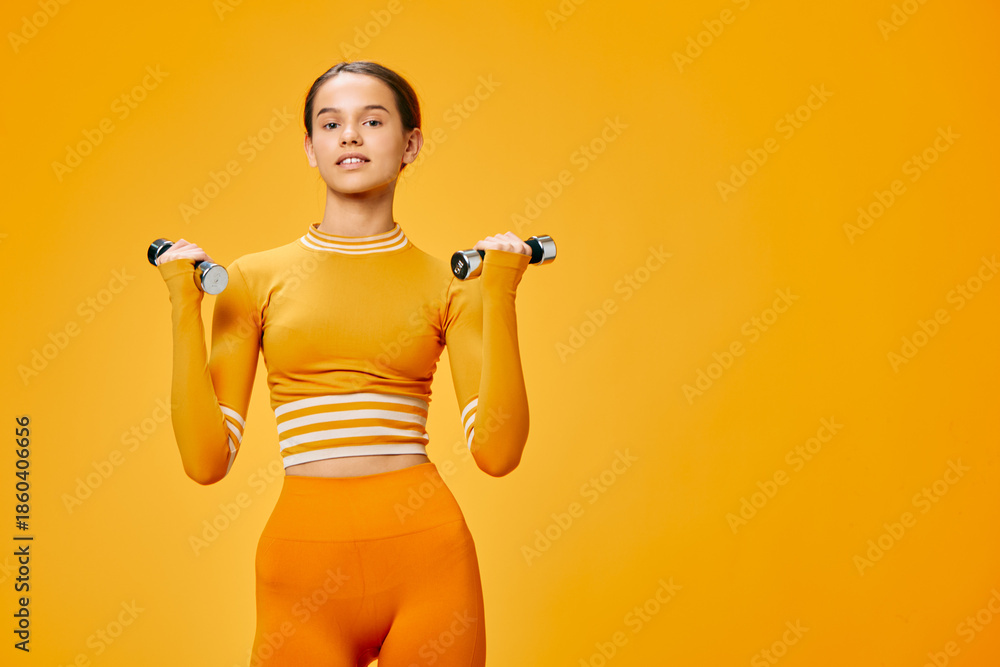 Fototapeta premium Woman fitness enthusiast with dumbbells in an orange studio, wearing a cropped top and leggings, demonstrating strength, balance and focus during a dynamic workout session.