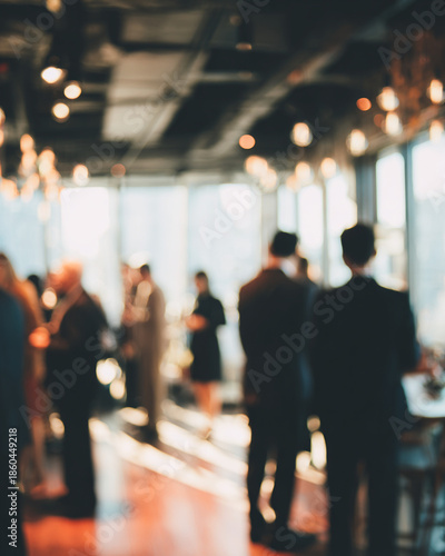 A group of people in business attire are socializing and standing in a large room with large windows and warm lighting.