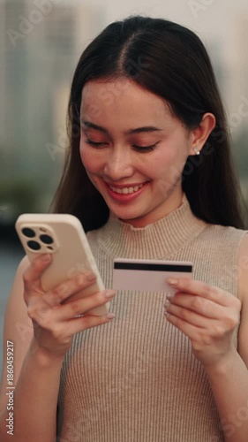 Portrait of pretty Asian woman enjoys online shopping with credit card and smartphone in city vertical shot. Young Japanese lady pays via banking app to protect money against park