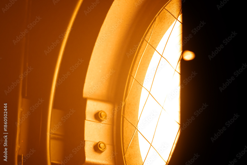 Obraz premium Close up of a glowing orange stage spotlight showing metal parts, bolts, and protective wire mesh. Lighting equipment backstage.
