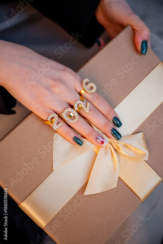 Female hand wearing shiny number rings forming 2026 delicately holds a brown gift box with golden ribbon and bow, festive close-up evoking holiday celebration and luxury joy