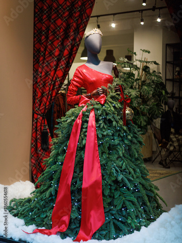 Holiday shop window display with a mannequin wearing a dress made of evergreen branches and red ribbon. Concept of creative retail decoration for Christmas branding, seasonal marketing.