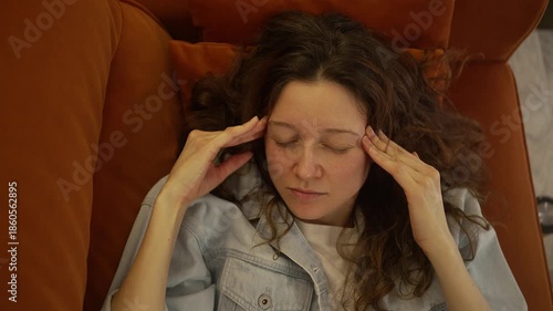 Overhead shot of a young woman dealing with a severe headache, resting on the sofa and gently rubbing her temples. An exhausted individual feeling unwell, enduring the discomfort of a migraine