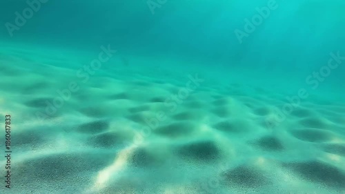 Underwater scene showing sandy ocean floor with gentle waves and sunlight filtering through the water