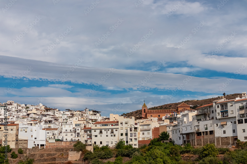Obraz premium A panoramic view of Torrox Pueblo displays the dense white houses and the church tower against a blue sky. The traditional architecture of this Andalusian town rests on the hills of the Costa del Sol.