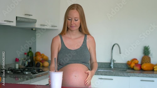 Pregnant woman stands in kitchen holding ice cream, wondering if it's safe for her and the baby. Concept of pregnancy cravings, nutrition awareness, and balancing health with joy. Slowmotion video.