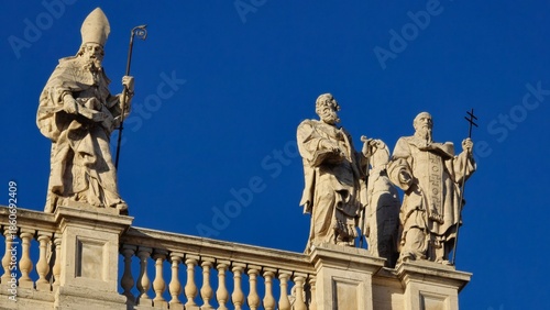 Rome, Italy - 14 January 2025. Rooftop statues on San Giovanni in Laterano depict a bishop and two saints with a staff, book, and cross above a balustrade under clear skies.