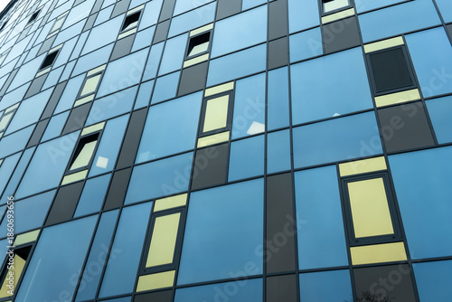 Modern building facade with blue and yellow glass windows in geometric grid pattern. Abstract urban architecture with reflective blue panels and asymmetric open windows on a contemporary glass facade.