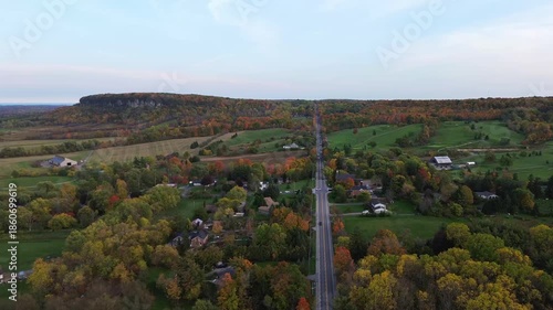4K top-down cinematic drone shot flying above forest and farmland alongside forest path road carved through colorful maple trees of autumn, with city downtown skyline in view, Ontario Canada
