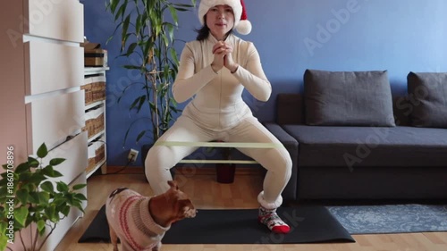 A young Caucasian woman wearing a Santa hat and Christmas socks performs squats with an elastic band on a mat while a dog in a sweater watches. Medium shot, slow motion, side view. Home fitness concep