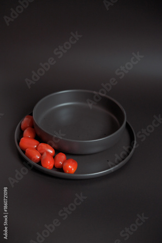 Shiny red plum tomatoes arranged on a matte black plate, with a matching black bowl stacked behind. Minimalist design emphasizing color contrast.