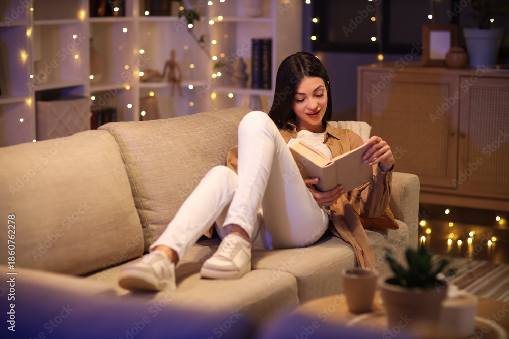 Obraz premium Beautiful young woman reading book and sitting on sofa in living room in evening
