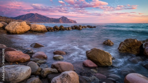 Waves gently wash over a rocky shore as the sun sets in the background. Mountains stand tall against a colorful sky filled with clouds. This scene captures nature's beauty at twilight.