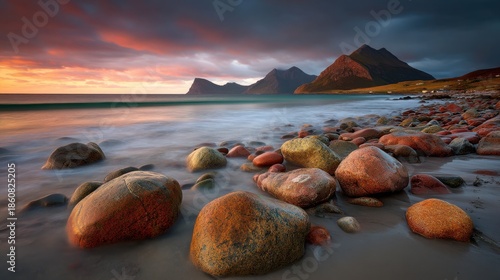 Brightly colored stones cover a beach at sunset. The sky shows shades of orange and purple. Mountains rise in the background. Waves gently hit the shore.