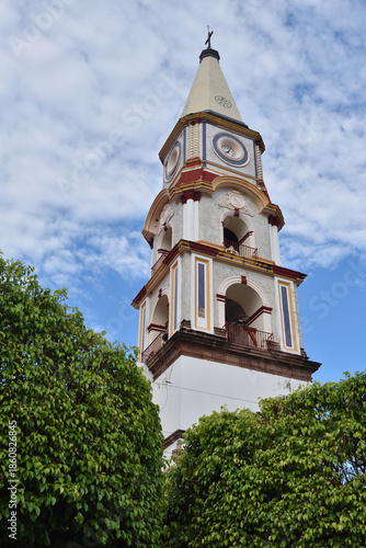 Vertical view of the ornate bell tower of the historic church in Mascota, Jalisco, rising above lush green trees under a bright blue sky
