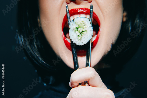 Close-up of a woman holding sushi with chopsticks, showcasing vibrant red lips and a playful expression against a dark background.