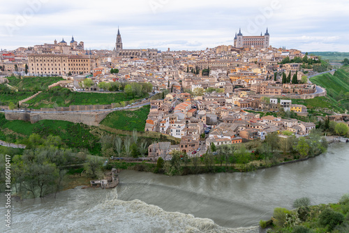 The ancient city of Toledo Spain glows softly at spring twilight with the Tagus River curving around the historic walls forming a natural barrier while lush greenery frames this elevated view.