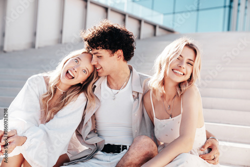 Young three stylish friends posing in the street. Fashion man and two cute female dressed in casual summer clothes. Smiling models having fun. Cheerful women and guy outdoors, in sunny day, sunglasses