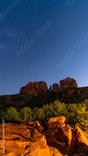 A nighttime panorama showcases a rock formation silhouette beneath a starry sky. Dark, blurred foreground leads to a forest line at the base