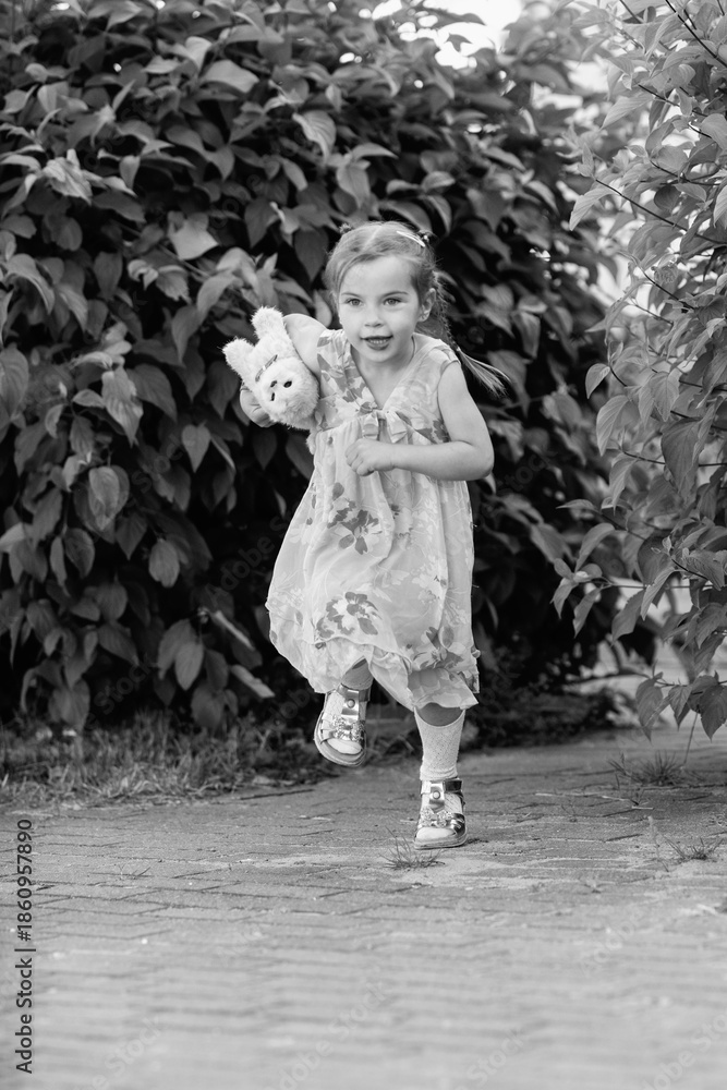 Naklejka premium Young girl with a teddy bear running joyfully across a grassy field, wearing a light dress and sneakers, capturing the essence of childhood playfulness and freedom