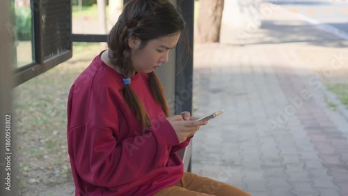 Asian woman sitting at bus stop using smartphone seriously during daytime urban lifestyle