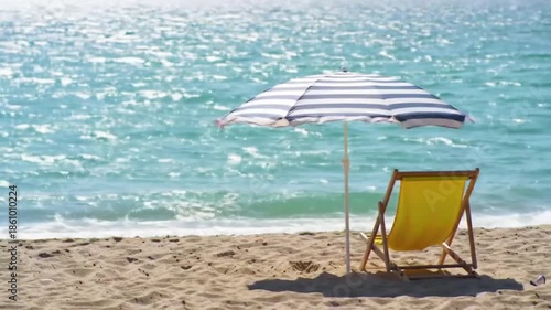 A solitary yellow lounge chair sits under a striped umbrella on a sandy beach, with sparkling blue ocean waves gently lapping the shore.