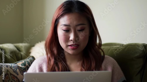 A young woman with red-toned hair sits on a green couch, looking attentively at a laptop. The background is softly lit