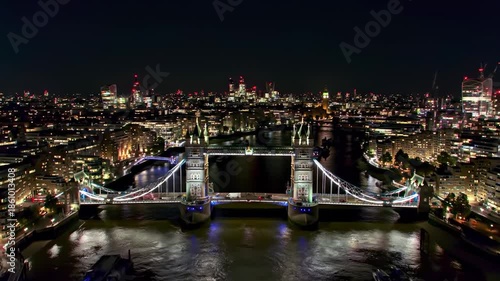 Aerial night view of a historic bridge, illuminated with blue lights, spanning a river. A cityscape of lights surrounds