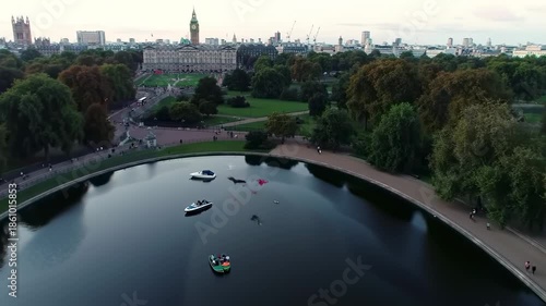 Aerial view of a city park with a lake, boats, and a prominent building with a tall clock tower in the background, surrounded by trees