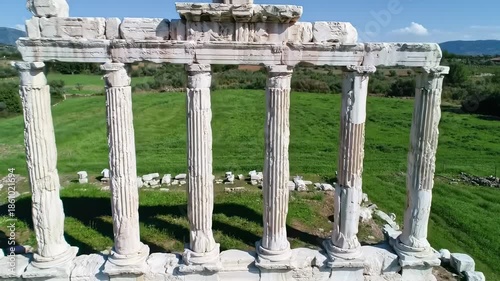 Aerial view of ancient colonnade in ruins with six fluted pillars, standing on a stone base, framing a grassy field with trees