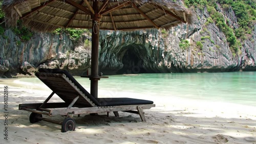 Serene beach lounge chair awaits under a thatched umbrella with a tropical cave in the background.