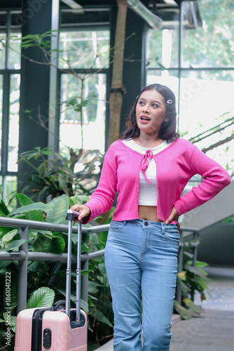 Young woman pulling her suitcase in an outdoor space brimming with verdant plants. She shows an expression of surprise as she begins an eagerly anticipated trip.