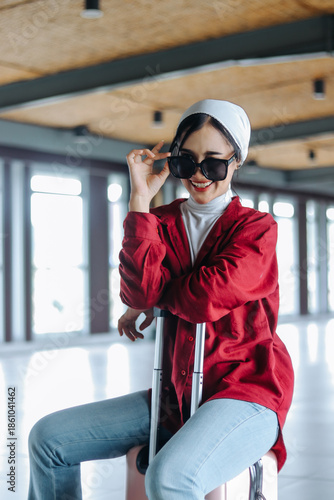 Attractive young woman sits with confidence atop her suitcase within the airy atmosphere of an airport. She's adorned in stylish shades, ready for her upcoming explorations.