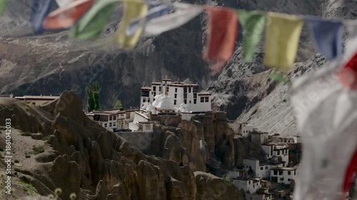 Tibetan gompa nestled on rocky mountainous landscape with slow motion colourful prayer flags fluttering in light breeze.
