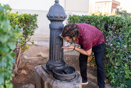 Man with dreadlocks drinking water from a traditional public fountain in the city