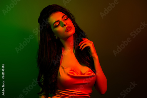 Glamorous young woman with dark hair in satin blouse illuminated by neon green and red light in artistic background